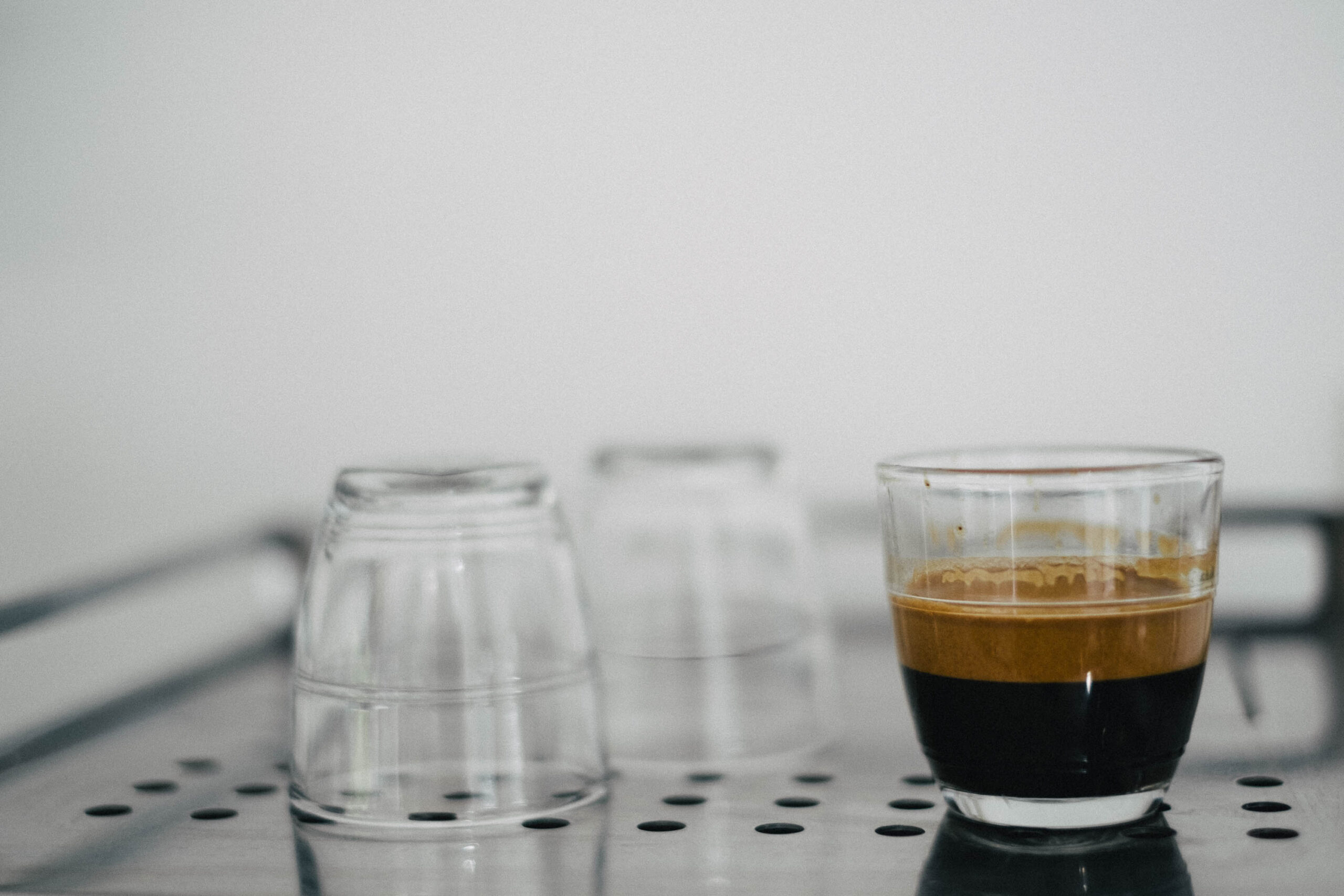 three small glass espresso cups, sitting on the metal top of a coffee machine (not pictured). two are empty, placed facing downwards, the third is 2/3 full of espresso coffee.